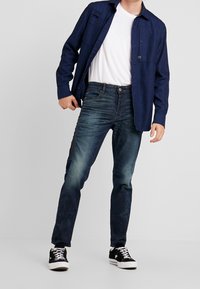 Marineblauw overhemd over een wit T-shirt, gecombineerd met blauwe denim jeans en zwarte sneakers met witte zolen; casual outfit, zonder zichtbare patronen.