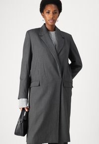 Blazer oversize grigio a righe sottili con revers a punta, due tasche frontali e una vestibilità sagomata, abbinato a una borsa nera e a un maglione in maglia.