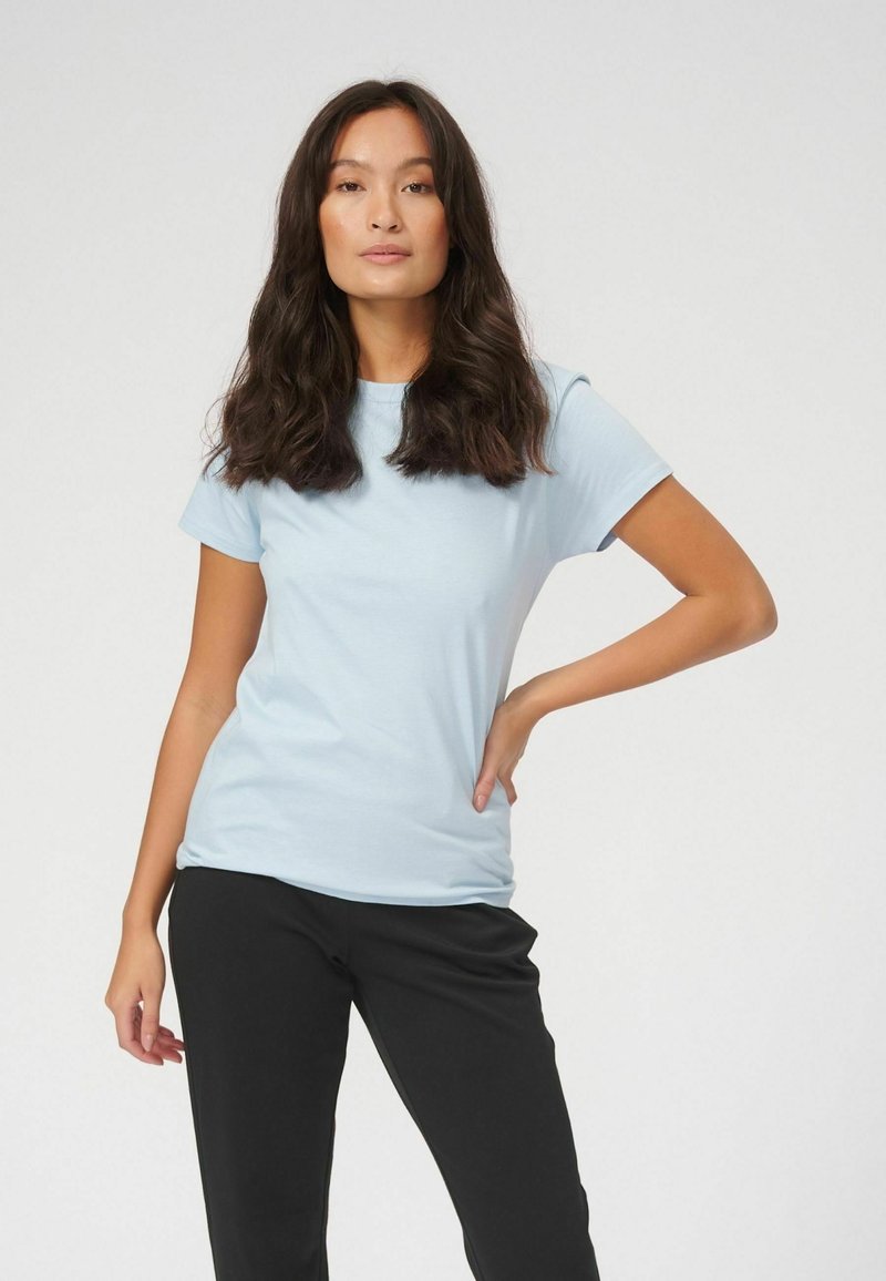 Femme aux longs cheveux foncés portant un t-shirt bleu clair et un pantalon noir, debout avec une main sur la hanche devant un fond uni.