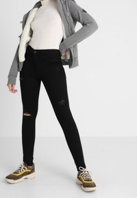 Šedá mikina s beránkovým podšívkou, bílý rolák, černé skinny džíny s trhanými detaily a hnědé/šedé tenisky se žlutými akcenty.