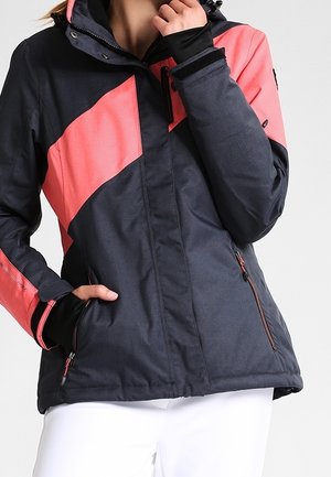 Nainen, joka pukeutunut tummanharmaaseen ja korallinväriseen talvitakkiin, jossa on vetoketjulliset taskut ja peukalonreiät hihoissa, poseeraa valkoista taustaa vasten.