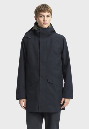 Chaqueta impermeable en color azul marino con capucha, cierre frontal de botones a presión y dos grandes bolsillos, diseñada con un corte holgado y una textura suave.