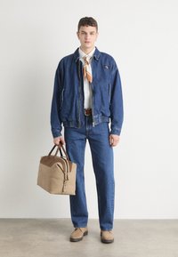 Veste en denim et jeans, chaussures brunes, avec un sac beige à poignées marron foncé. Chemise blanche et écharpe orange à motifs.