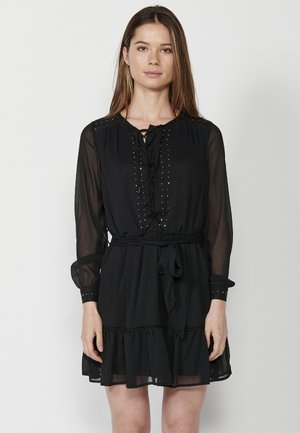 Schwarzes Kleid mit einem gerafften Saum, Spitzenverzierungen und langen durchsichtigen Ärmeln. Weicher Stoff mit einem Bindeband in der Taille und dekorativen Nieten entlang des Oberteils.