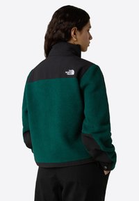 Giacca in pile di un verde scuro con accenti neri, colletto alto e tessuto strutturato. Presenta un logo sulla spalla posteriore.