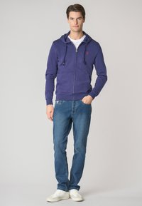 Felpa con zip di colore viola, con cappuccio regolabile, tasche frontali e logo rosso. Abbinata a jeans azzurri chiari e scarpe da ginnastica bianche.