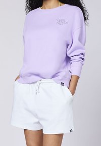 Mujer con sudadera lila con el texto “SOLO BUENAS OLAS” y pantalones cortos blancos con cordones, con las manos en los bolsillos, cabello rizado visible.