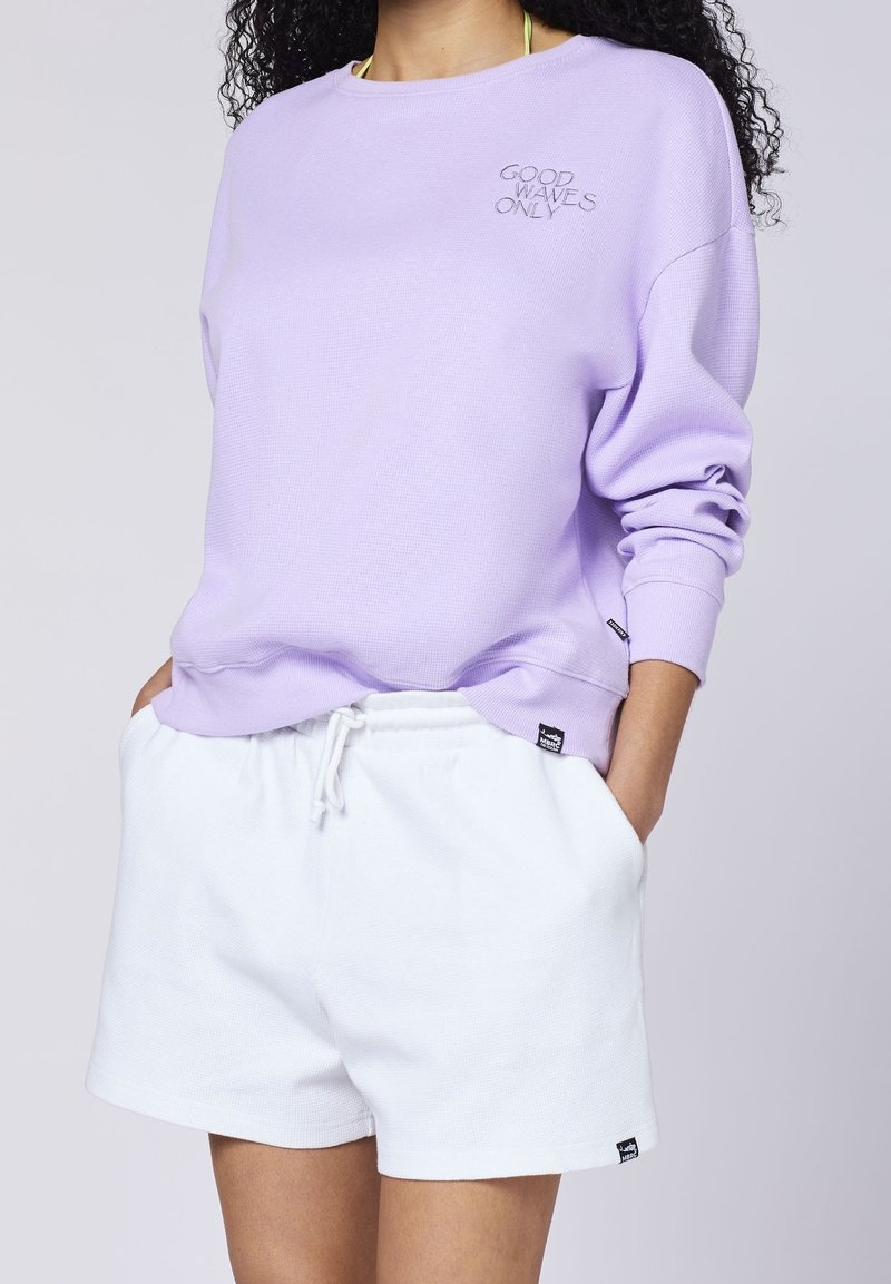 Mujer con sudadera lila con el texto “SOLO BUENAS OLAS” y pantalones cortos blancos con cordones, con las manos en los bolsillos, cabello rizado visible.