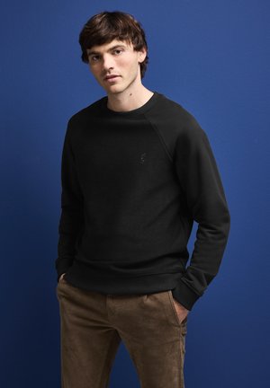 Sweatshirt noir avec un col rond, des poignets et un ourlet côtelés ; porté sur un pantalon en velours côtelé marron. L'arrière-plan est d'un bleu uni.