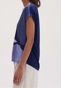 Kvinna som bär en kortärmad marinblå och lavendel geometriskt mönstrad topp med ljusa byxor, visad från sidan vid midjan.