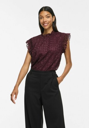 Femme souriante portant un chemisier sans manches texturé violet foncé avec des bords volantés et un pantalon noir à jambes larges taille haute, main dans la poche.