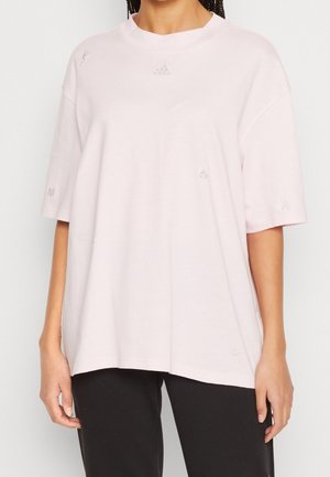 Femme portant un t-shirt Adidas rose clair ample avec des logos brodés subtils et un pantalon noir, debout devant un fond uni.