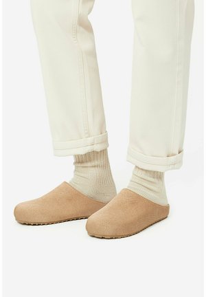 Stopy w beżowych kapciach wsuwanych, z jasnobeżowymi prążkowanymi skarpetkami i podwiniętymi jasnobeżowymi spodniami na białym tle.