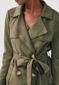 Manteau trench en faux daim vert olive avec une taille ceinturée, un design à double boutonnage, grands revers et deux poches frontales.