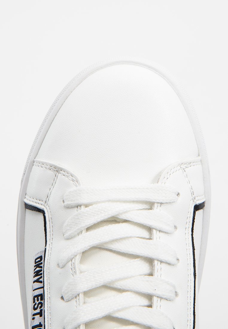 Witte synthetische sneaker met platte witte veters, gestikte details en een zwarte merkaccent aan de zijkant. Gladde textuur met ronde neus.