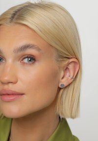 Strieborné náušnice na štuplíky s textúrovaným povrchom, sprevádzané tenkým strieborným kruhovým náušnicom. Vlasy sú upravené do rovného, blond bobu.