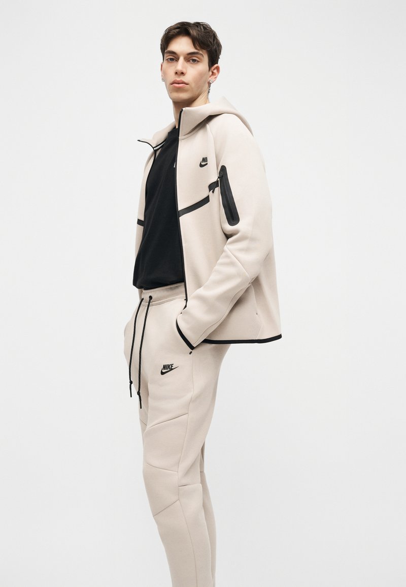 Joven con chándal beige de Nike con detalles en negro, de pie con las manos en los bolsillos contra un fondo blanco liso.