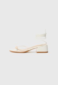 White heeled sandals with a square heel, thin straps, and a round accent. Features a textured sole and a coiled lace detail at the ankle.