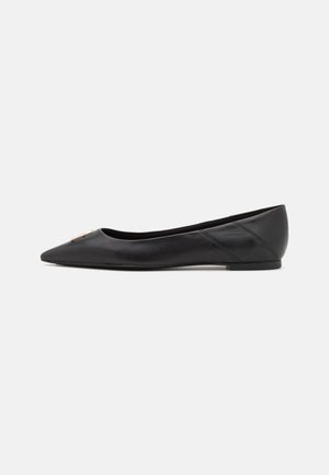 Chaussure plate en cuir noir à bout pointu avec petit talon et petit emblème décoratif doré près de la pointe, vue de côté sur fond clair uni.