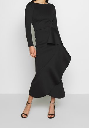Vrouw draagt een lange zwarte jurk met lange mouwen en een asymmetrische gerimpelde rok, gecombineerd met zwarte enkellaarzen met riempjes.