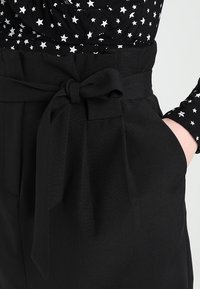 Svarta högmidjade byxor med ett knytbart tygbälte. Har sidofickor och en texturerad tyg. Kombineras med en svart topp med vitt stjärnmönster.