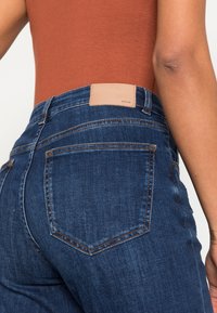 Tmavě modré džíny z denimu mají přiléhavý střih s hladkou strukturou a kontrastním oranžovým prošíváním. Na zadní straně je viditelná světle hnědá kožená nášivka.