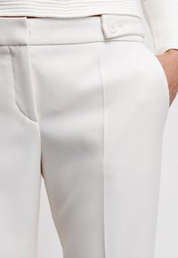 Close-up van witte op maat gemaakte broek met een knoopsluiting en een hand in de zak, gecombineerd met een textuur wit top.