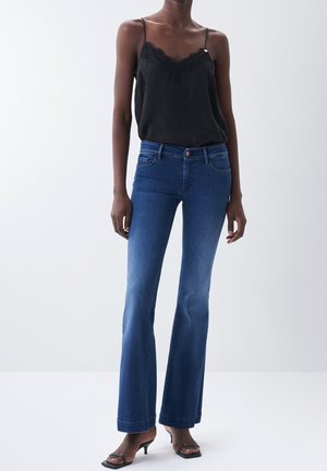 Kvinde iført sort ærmeløs top med blonder, blå flared jeans og sorte åbne kitten heels foran en ensartet lys baggrund.