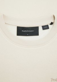 Felpa beige chiaro con colletto e polsini a coste. Presenta un'etichetta nera con la scritta "PeakPerformance" e "Made in Turkey". Taglia media.