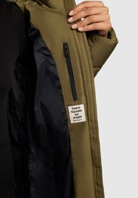 Jachetă puffer de culoare verde măslin, cu interior căptușit în negru. Dispune de un buzunar lateral cu fermoar și o etichetă care spune "Un îmbrățișare călduroasă din țesătură."