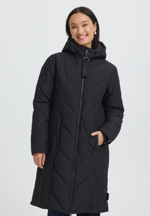 Femme souriante portant un long manteau d'hiver noir matelassé avec capuche, debout avec une main dans la poche devant un fond uni.