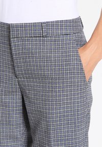 Person klädd i grå och blå rutiga byxor med en hand delvis i den högra framfickan, i kombination med en vit topp.