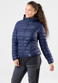 Navy puffer jacket with a hood, featuring a quilted design, front zipper, and logo. Paired with dark pants with zippered pockets.