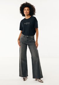 Frau mit lockigem Haar, die ein schwarzes Grafik-T-Shirt, hochgeschnittene, weit geschnittene graue Jeans und metallische, spitze Absatzschuhe vor einem weißen Hintergrund trägt.