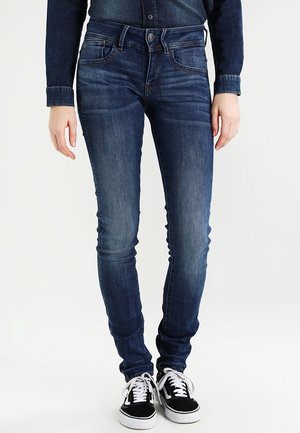 Tmavě modré džíny slim střihu nošené s černo-bílými teniskami a odpovídající tmavou denimovou košilí na stojící osobě.