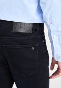 Jean en denim bleu foncé à texture lisse, avec un patch en cuir noir embossé du mot "BOSS" et un détail logo en métal sur la poche.