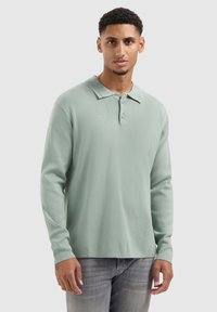 Helles grünes Langarm-Polohemd mit weicher Textur, ausgestattet mit einem spitzen Kragen und einer dreiknopflangen Knopfleiste, kombiniert mit grauen Jeans.