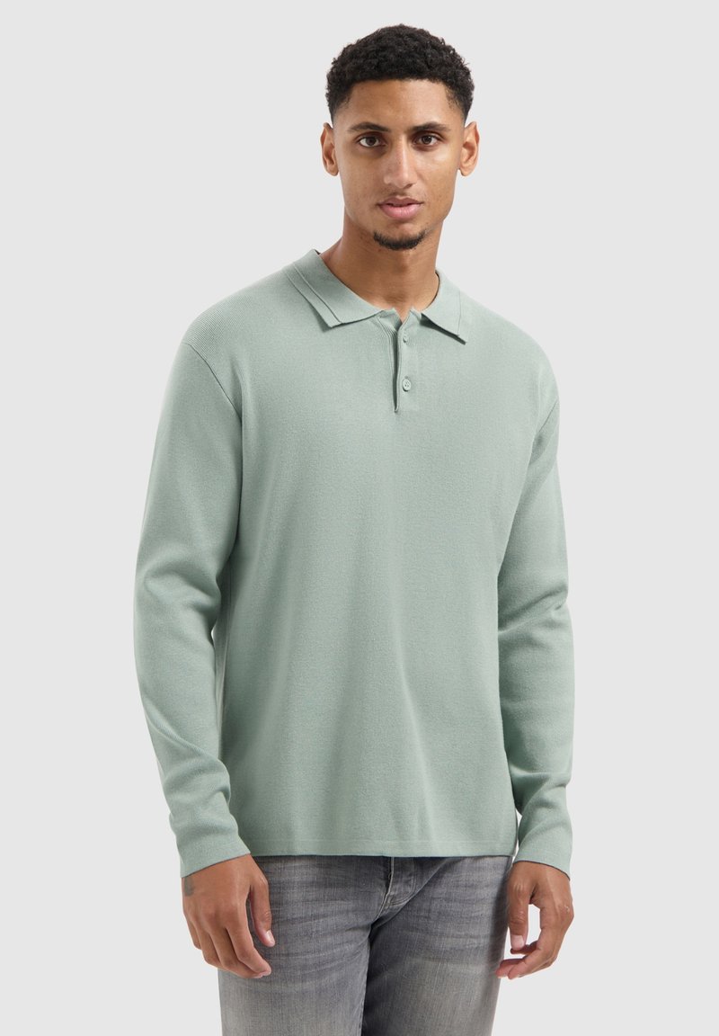 Helles grünes Langarm-Polohemd mit weicher Textur, ausgestattet mit einem spitzen Kragen und einer dreiknopflangen Knopfleiste, kombiniert mit grauen Jeans.