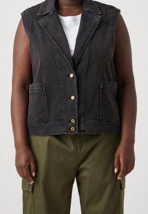 Zwarte mouwloze spijkervest met revers en gouden knopen, gedragen over een wit shirt, gecombineerd met olijfgroene cargobroek.