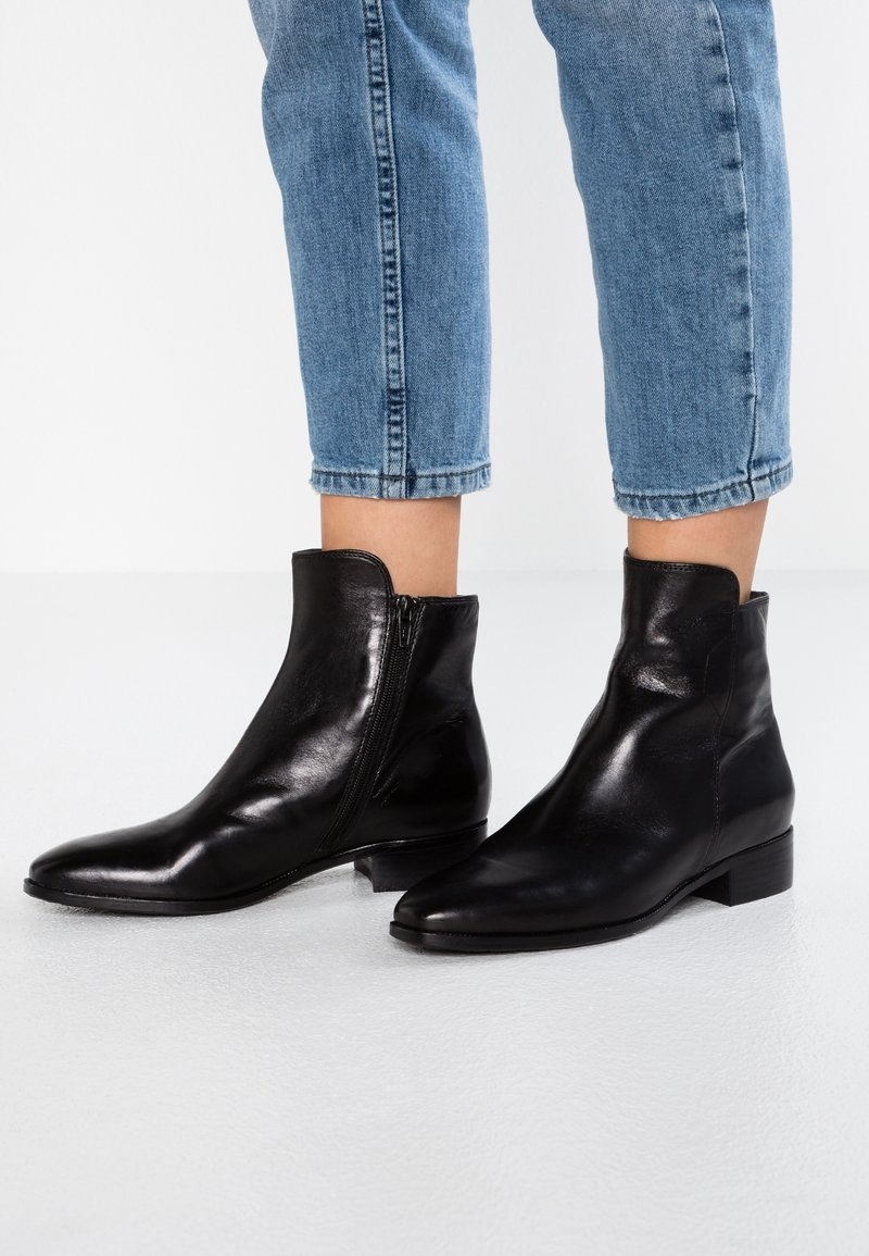 Personne portant des bottines en cuir noir mi-mollet avec des fermetures éclair sur le côté et un jean bleu foncé remonté contre un fond blanc uni.