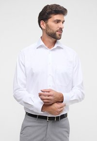 Witte lange mouwen shirt met een button-down kraag, een enkele borstzak en een gladde textuur. Gecombineerd met grijze broek en een zwarte riem.