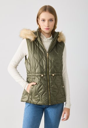 Quilted olive green vest with faux fur hood, gold zipper, drawstring waist, and side pockets, worn over a white long-sleeve top.