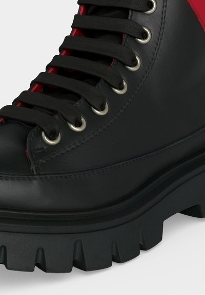 Botte en cuir noir avec semelle épaisse à crampons, lacets noirs passant à travers des œillets métalliques et doublure rouge visible sous les lacets.