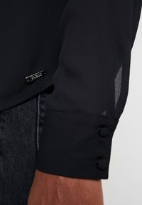 Blouse noire avec panneaux de manches translucides, boutons décoratifs aux poignets, et une petite étiquette métallique de la marque à l'ourlet. Texture lisse.