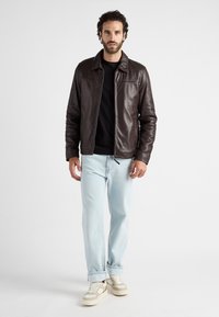 Braune Lederjacke mit glatter Textur, ausgestattet mit einem Kragen und subtilen Nähten. Kombiniert mit hellblauen Jeans und cremefarbenen Sneakers.