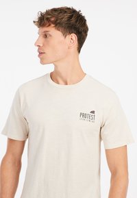 Lys beige kortærmet t-shirt lavet af blødt stof, med et svart logo på venstre bryst og en rund halsudskæring.