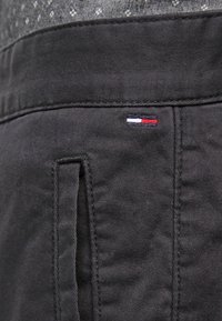 Pantalon en tissu noir avec une poche latérale verticale et un petit patch logo rectangulaire rouge, blanc et bleu près de la ceinture.