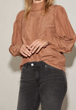 Vrouw draagt een roestkleurige blouse met hoge hals, pofmouwen en manchetten met knopen, gecombineerd met donkergrijze spijkerbroek.