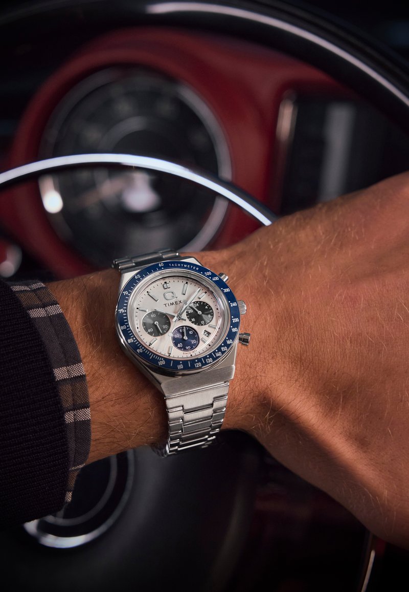 Montre en acier inoxydable argentée avec une lunette bleue, un cadran blanc et des sous-cadrans foncés, portée sur un poignet vêtu d'une chemise à carreaux.