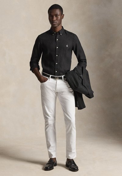 Άνδρας που φοράει μαύρο πουκάμισο με κουμπιά, λευκό παντελόνι, μαύρα μοκασίνια, κρατώντας ένα μαύρο σακάκι με το ένα χέρι στην τσέπη, ουδέτερο φόντο.